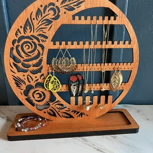 Peut inclure: Un présentoir à bijoux en bois de forme circulaire avec des découpes florales. Le présentoir comporte plusieurs fentes pour boucles d'oreilles et colliers. Un bracelet et divers boucles d'oreilles et colliers sont exposés. Le bois est brun chaud.