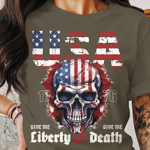 May include: A dark green t-shirt featuring a skull graphic and the text "USA 1776 Give Me Liberty or Death". The skull is designed with the colours and pattern of the American flag.