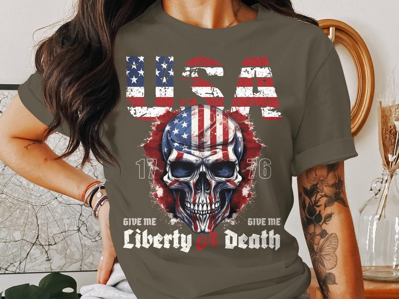 May include: A dark green t-shirt featuring a skull graphic and the text "USA 1776 Give Me Liberty or Death". The skull is designed with the colours and pattern of the American flag.