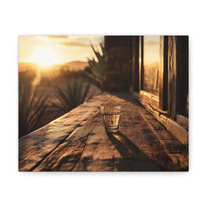 May include: A glass of clear liquid sits on a wooden surface with a blurred background of a sunset and agave plants.