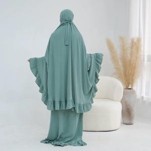 May include: A sage green prayer garment featuring a hooded head covering with tie closures and a long, flowing silhouette. The piece includes ruffled trim along the edges of the sleeves and the bottom hem, creating a layered, textured appearance.