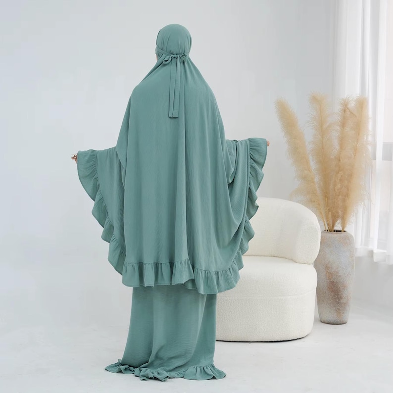 May include: A sage green prayer garment featuring a hooded head covering with tie closures and a long, flowing silhouette. The piece includes ruffled trim along the edges of the sleeves and the bottom hem, creating a layered, textured appearance.