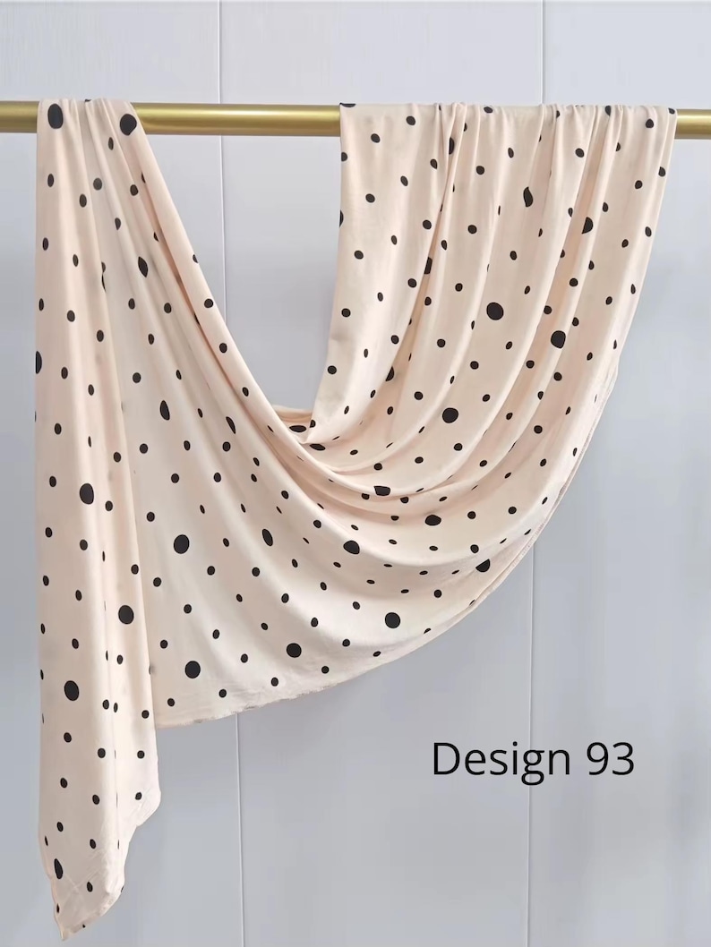 May include: A light beige fabric featuring a pattern of black polka dots of varying sizes, draped over a horizontal gold rod. The fabric hangs in soft folds, and the text Design 93 is printed in black in the lower right corner of the image.
