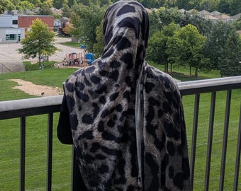 May include: A headscarf featuring a black and gray snow leopard print pattern. The fabric drapes over the head and shoulders, displaying a repeating animal spot design. The text "Snow Leopard Print" is clearly visible at the top of the image.