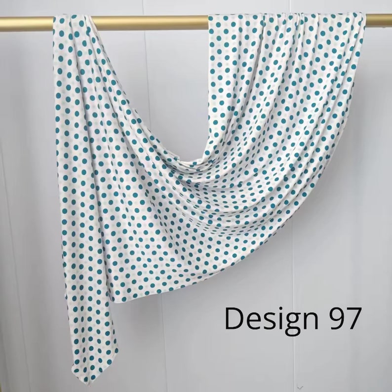 May include: A white fabric scarf featuring a repeating pattern of teal and light gray polka dots, draped over a gold rod. The fabric is labeled as Design 97, showcasing a soft, flowing texture with consistent circular motifs across the material.