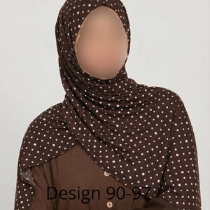 May include: A dark brown hijab featuring a white polka dot pattern draped over the head and shoulders. The fabric is layered around the neck and chest area. A solid dark brown shirt with light-colored buttons is visible underneath. Text reads Design 90-97.