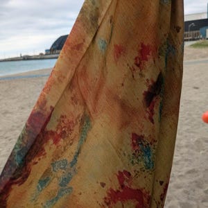 May include: A draped scarf with a watercolour design. The fabric displays a mix of warm colours, such as yellow, orange, and red, with splashes of blue. The scarf's texture and pattern are visible against a beach background.