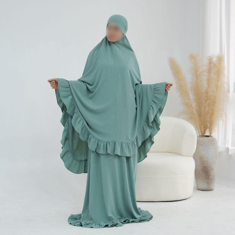 May include: A sage green prayer dress featuring a matching head covering and a long, flowing silhouette. The garment is accented with ruffled trim along the edges of the sleeves and the bottom hem, providing a layered look in a solid color.