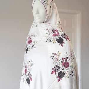 May include: A white hijab featuring a floral print with dark red and black roses, green leaves, and thin stems. The head covering is draped over a mannequin head and shoulders, displaying a repeating pattern of botanical elements across the fabric.