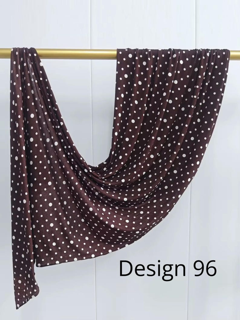 May include: A dark brown rectangular scarf featuring a repeating pattern of small white polka dots. The fabric is draped over a horizontal gold rod, creating soft folds and ripples. The text Design 96 is printed in black in the lower right corner.