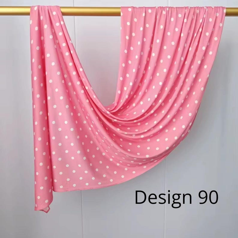 May include: A pink fabric featuring a repeating white polka dot pattern draped over a gold rod. The fabric hangs in soft, vertical folds, displaying a consistent dotted design throughout. The text "Design 90" is printed in black in the lower right.