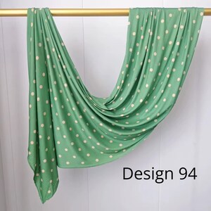 May include: A sage green fabric scarf featuring a repeating pattern of small, cream-colored polka dots. The material is draped over a horizontal gold rod, creating soft folds and ripples. The text "Design 94" is printed in black in the corner.