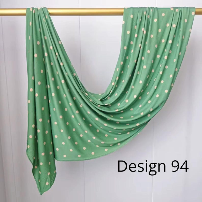 May include: A sage green fabric scarf featuring a repeating pattern of small, cream-colored polka dots. The material is draped over a horizontal gold rod, creating soft folds and ripples. The text "Design 94" is printed in black in the corner.