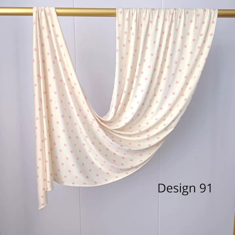 May include: A cream-colored fabric draped over a gold rod, featuring a repeating pattern of small, light pink polka dots. The fabric hangs in soft, vertical folds, and the text "Design 91" is printed in black in the lower right corner.
