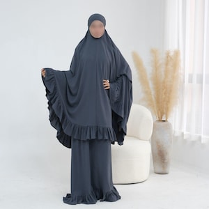 May include: A dark charcoal gray two-piece prayer garment consisting of a long, loose-fitting khimar with a ruffled hemline and matching wide-leg trousers that also feature a ruffled bottom edge, providing full coverage in a solid, matte fabric.