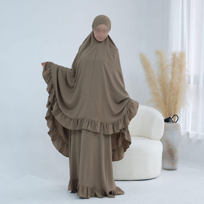 May include: A taupe-colored two-piece prayer garment consisting of a long, loose-fitting head covering with a ruffled hem and a matching floor-length skirt with a ruffled bottom edge. The set features a solid, matte fabric in a neutral earth tone.