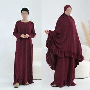 May include: A deep burgundy long-sleeved maxi dress with a ruffled hemline, paired with a matching burgundy head covering that features a ruffled edge. The outfit is shown in two views, one with a black head cap and one with the full head covering.