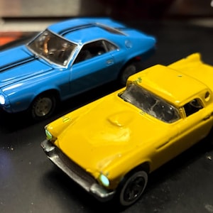Peut inclure: Deux voitures miniatures : une voiture de sport bleu vif avec des rayures noires et une voiture vintage jaune. La voiture bleue a un pare-brise argenté et la voiture jaune a des pneus à flancs blancs. Les deux sont sur une surface sombre.