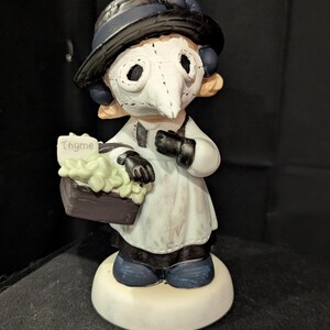 May include: A ceramic figurine of a plague doctor wearing a black hat and mask, holding a basket of thyme. The figurine is dressed in a white coat and black gloves, standing on a white base.