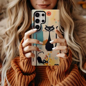 May include: A rectangular smartphone case featuring a cream-colored background with two black cat illustrations, blue geometric shapes, and orange circular accents. The design includes small starburst patterns and a multi-lens camera cutout at the top.