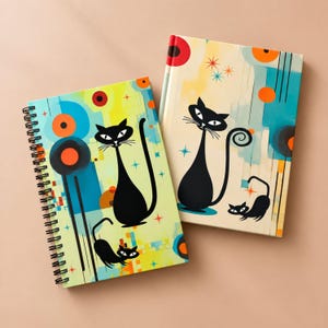 May include: Two notebooks featuring black cat illustrations with long tails and pointed ears. The covers display abstract geometric shapes, circles, and starburst patterns in shades of blue, orange, yellow, and cream with black spiral binding.