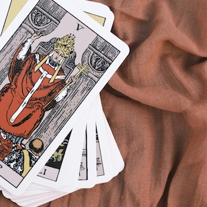 May include: A deck of tarot cards fanned out, featuring the Hierophant card prominently. The card displays a figure in a red robe and gold crown holding a triple cross staff, with the Roman numeral V at the top. The cards have white borders.