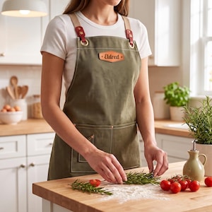 May include: An olive green canvas apron featuring a tan leather patch with the name Eldred embossed in black. The apron includes tan shoulder straps secured with red leather tabs and brass hardware, along with two front pockets and white stitching.