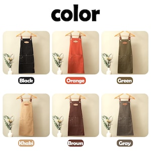 May include: A collection of six aprons displayed in black, orange, green, khaki, brown, and gray. Each apron features two front pockets, a leather-like chest patch, and adjustable straps with metal hardware, all hanging on wooden hangers.