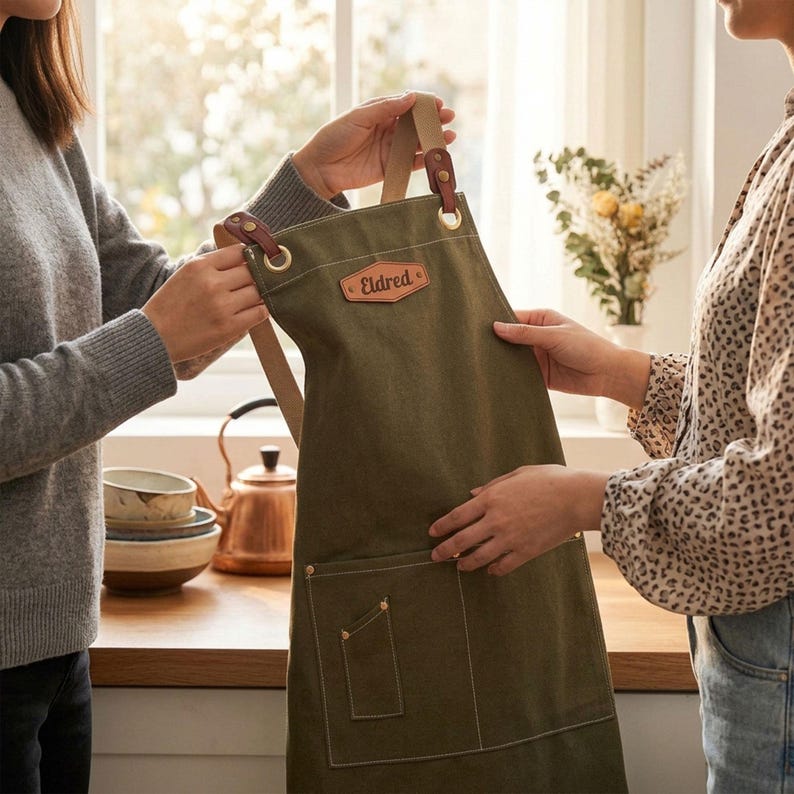 May include: An olive green canvas apron featuring a tan leather patch with the name Eldred embossed in brown. The apron includes tan fabric straps attached with brown leather tabs and brass-colored metal grommets, plus a large front pocket.