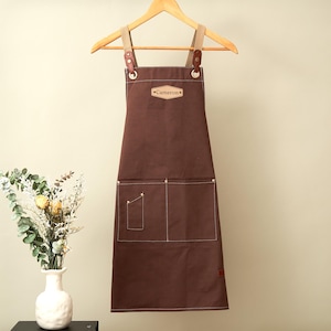 May include: A dark brown canvas apron featuring a light brown leather patch with the name Cameron embossed in the center. The apron includes two large front pockets, one smaller utility pocket, and adjustable straps attached with metal hardware.