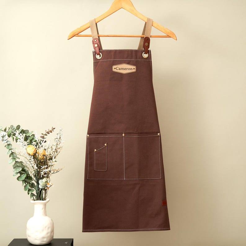 May include: A dark brown canvas apron featuring a light brown leather patch with the name Cameron embossed in the center. The apron includes two large front pockets, one smaller utility pocket, and adjustable straps attached with metal hardware.