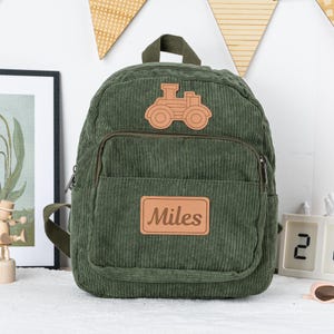 May include: A green corduroy backpack with a brown leather patch featuring a cartoon tractor and the name "Miles".