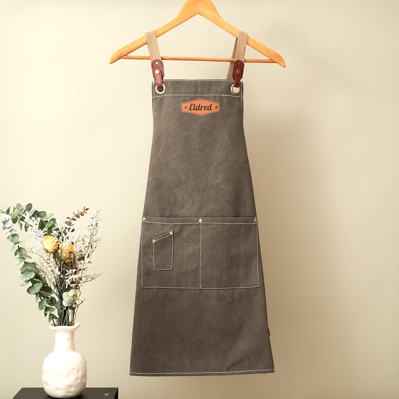 May include: A dark gray canvas apron featuring a leather patch with the name Eldred embossed on the chest. The apron includes two large front pockets, one small utility pocket, and adjustable tan straps secured with brown leather and metal hardware.