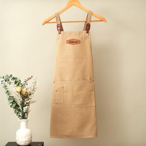 May include: A tan canvas apron featuring brown leather straps attached with metal rings and rivets. The apron includes a chest patch with the text Eldred, a large front pocket, and a smaller side pocket. It is displayed on a wooden hanger.