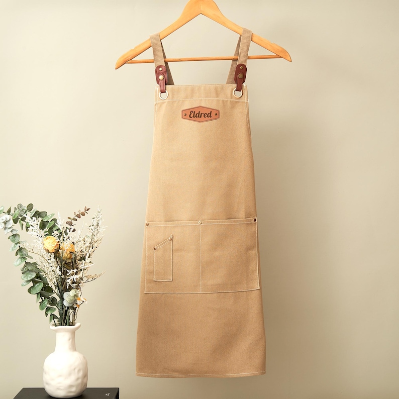 May include: A tan canvas apron featuring brown leather straps attached with metal rings and rivets. The apron includes a chest patch with the text Eldred, a large front pocket, and a smaller side pocket. It is displayed on a wooden hanger.