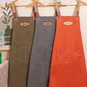 May include: Three personalized aprons in olive green, gray, and orange hang on wooden hangers. Each apron has a pocket and a leather tag with a name: "Liam", "Violet", and "William". The aprons have brown straps and metal hardware.