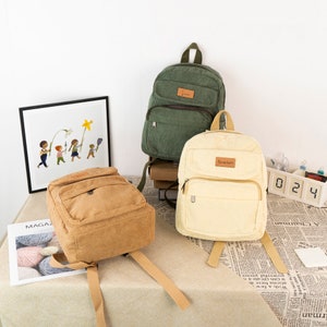 May include: Three corduroy backpacks in different colors: green, beige, and cream. The cream backpack has the word "Scarlett" on the front.