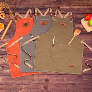May include: Three canvas aprons in orange, gray, and olive green, with leather accents and pockets. Each apron has a leather patch with a logo or name. A wooden spoon and a knife are in the pockets of the olive green apron.
