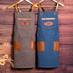 May include: Two aprons, one gray and one blue, hang on wooden hangers. Each apron has a brown leather pocket and a leather patch with a name. The aprons are made of a durable fabric, perfect for cooking or crafting.