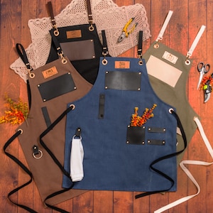 May include: A collection of aprons in various colors, including brown, black, blue, and olive green. Each apron features leather accents, metal grommets, and adjustable straps. Some aprons have pockets and a loop for a towel. The aprons are designed for utility and style.
