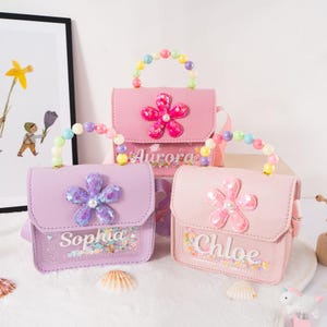 May include: Three pastel-colored purses with flower embellishments and beaded handles. The purses are pink, lavender, and light pink, each with a different name: "Aurora", "Sophia", and "Chloe". The purses have a clear panel with colorful confetti.