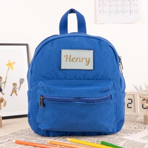 May include: A small blue canvas backpack with a zippered front pocket. The backpack has a name tag with the name "Henry" written in gold lettering.