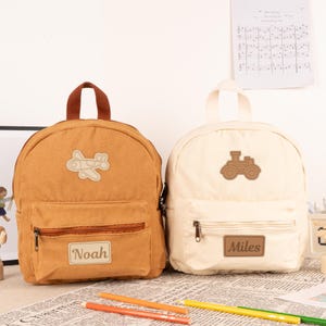 May include: Two personalised children's backpacks. One is brown with a plane patch and the name "Noah" on the front. The other is white with a toy truck patch and the name "Miles" on the front.