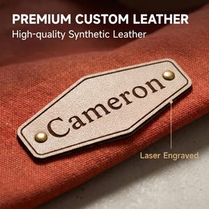 May include: A diamond-shaped beige synthetic leather patch with the laser-engraved name Cameron, secured by two brass-colored metal rivets onto a textured orange fabric. The text PREMIUM CUSTOM LEATHER, High-quality Synthetic Leather, and Laser Engraved appear.