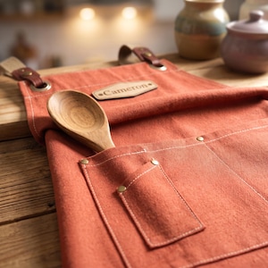 May include: A burnt orange canvas apron featuring a leather patch with the name Cameron embossed on it. The apron includes a large front pocket with brass rivets and a wooden spoon tucked inside. Brown leather straps are attached with metal grommets.