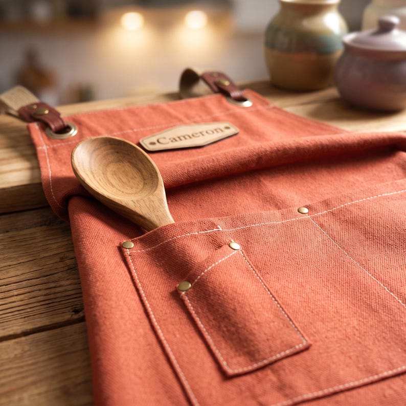 May include: A burnt orange canvas apron featuring a leather patch with the name Cameron embossed on it. The apron includes a large front pocket with brass rivets and a wooden spoon tucked inside. Brown leather straps are attached with metal grommets.