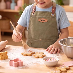 May include: An olive green apron with tan straps, brass-toned hardware, and a brown leather-like patch on the chest featuring the name Liam in black script. The apron includes two front pockets with copper-colored rivets at the corners.