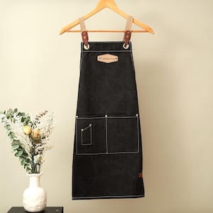 May include: A black apron featuring a tan leather patch with the name Cameron embossed on the chest. The apron has two large front pockets, one small side pocket, and tan fabric straps attached with brown leather tabs and metal rivets.