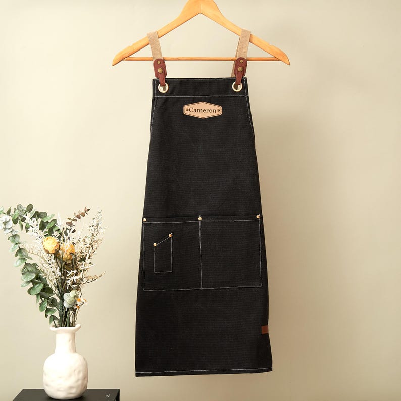 May include: A black apron featuring a tan leather patch with the name Cameron embossed on the chest. The apron has two large front pockets, one small side pocket, and tan fabric straps attached with brown leather tabs and metal rivets.
