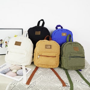 May include: Five mini backpacks in different colors and materials. The backpacks are made of canvas and have a zipper closure. The backpacks have different names on them, including "Leo" and "Jaime".